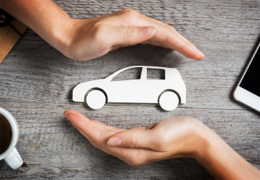 Deux mains protègent une voiture de papier déposée sur une table en bois.